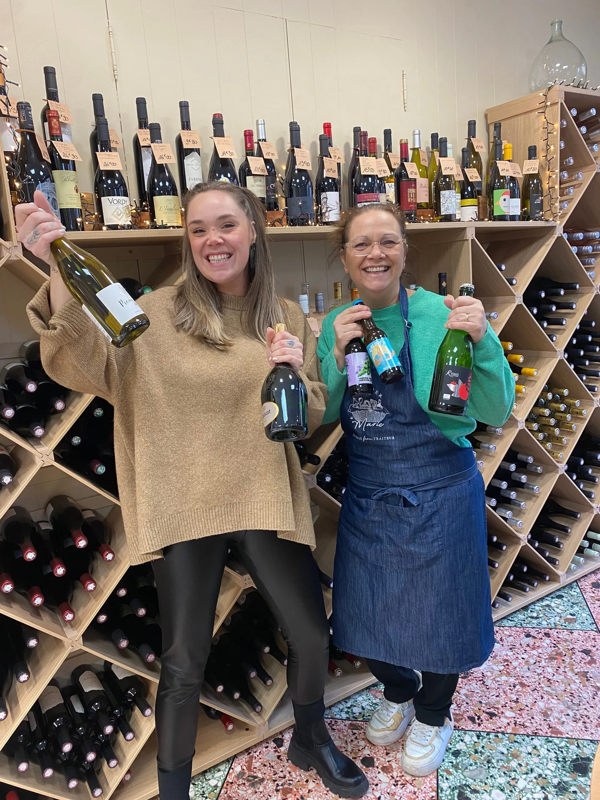 Marie et sa mère souriantes présentant des bouteilles de vin et de bière devant les casiers de la cave